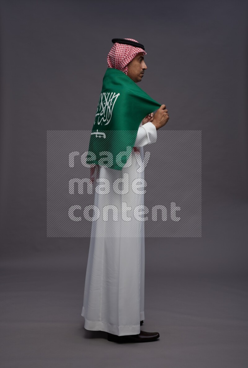 Saudi man wearing thob and shomag standing holding Saudi flag on gray background