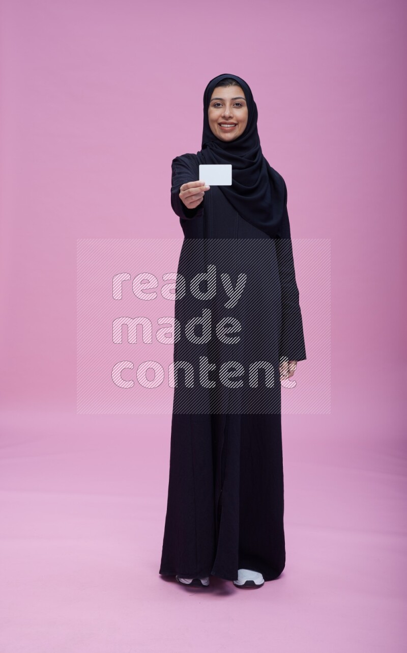 Saudi woman wearing Abaya standing holding ATM card on pink background