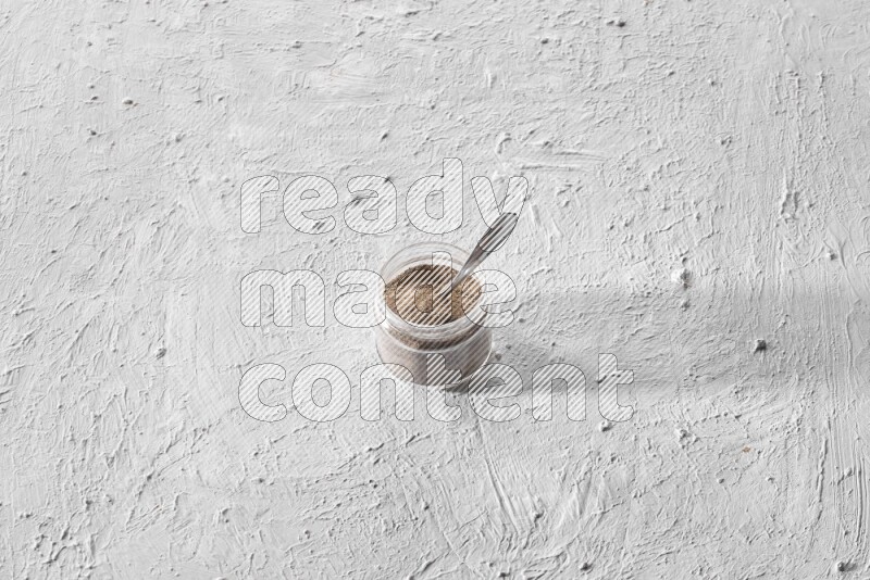 A glass jar full of black pepper powder and a metal spoon full of powder on a textured white flooring