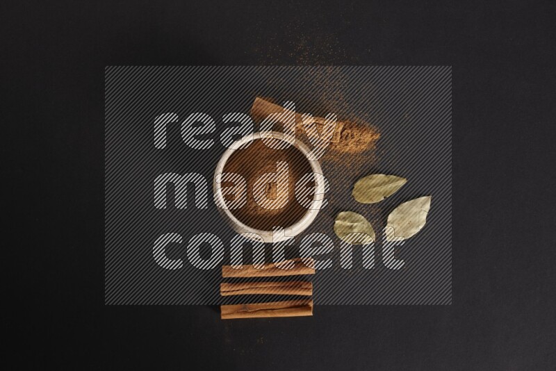 Cinnamon powder in a white pottery bowl and cinnamon sticks and laurel leaves on black background