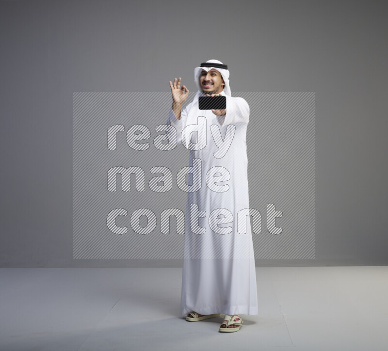 A Saudi man standing wearing thob and white shomag showing phone to camera on gray background