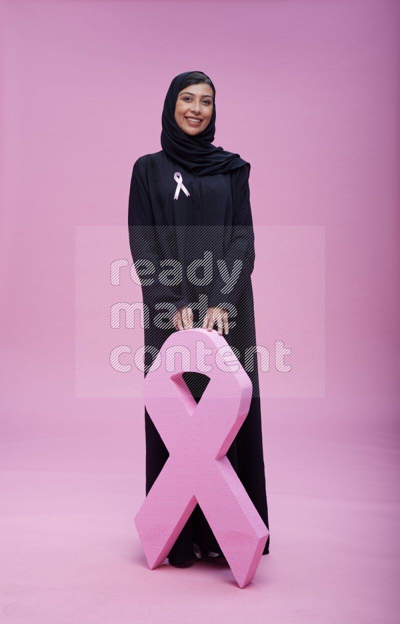 Saudi woman wearing pink ribbon on abaya standing holding awareness ribbon on plain pink background