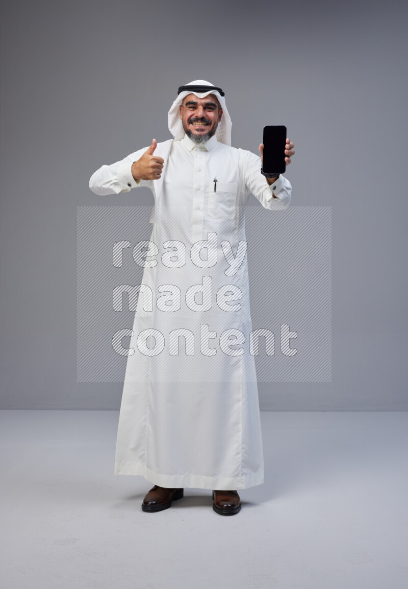Saudi man Wearing Thob and white Shomag standing showing phone to camera on Gray background
