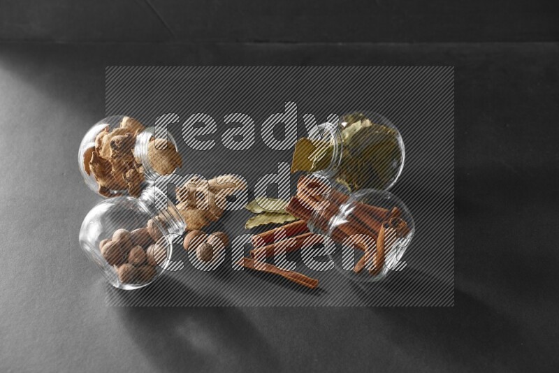 4 glass spice jars full of laurel bay leaves, ginger, cinnamon and nutmeg on black flooring