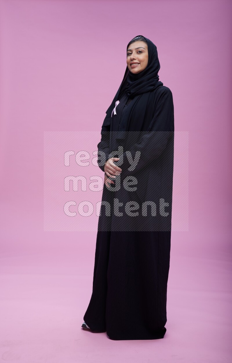 Saudi woman wearing pink ribbon on Abaya standing interacting with the camera on pink background
