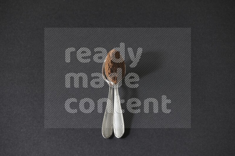 2 metal spoons on top of each other full of cloves powder on a black flooring