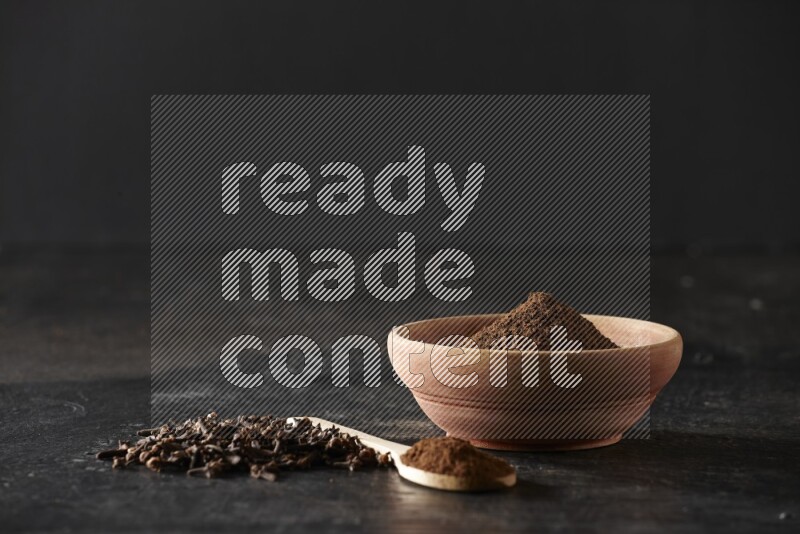 A wooden bowl and a wooden spoon full of cloves powder with spreaded cloves on a textured black flooring