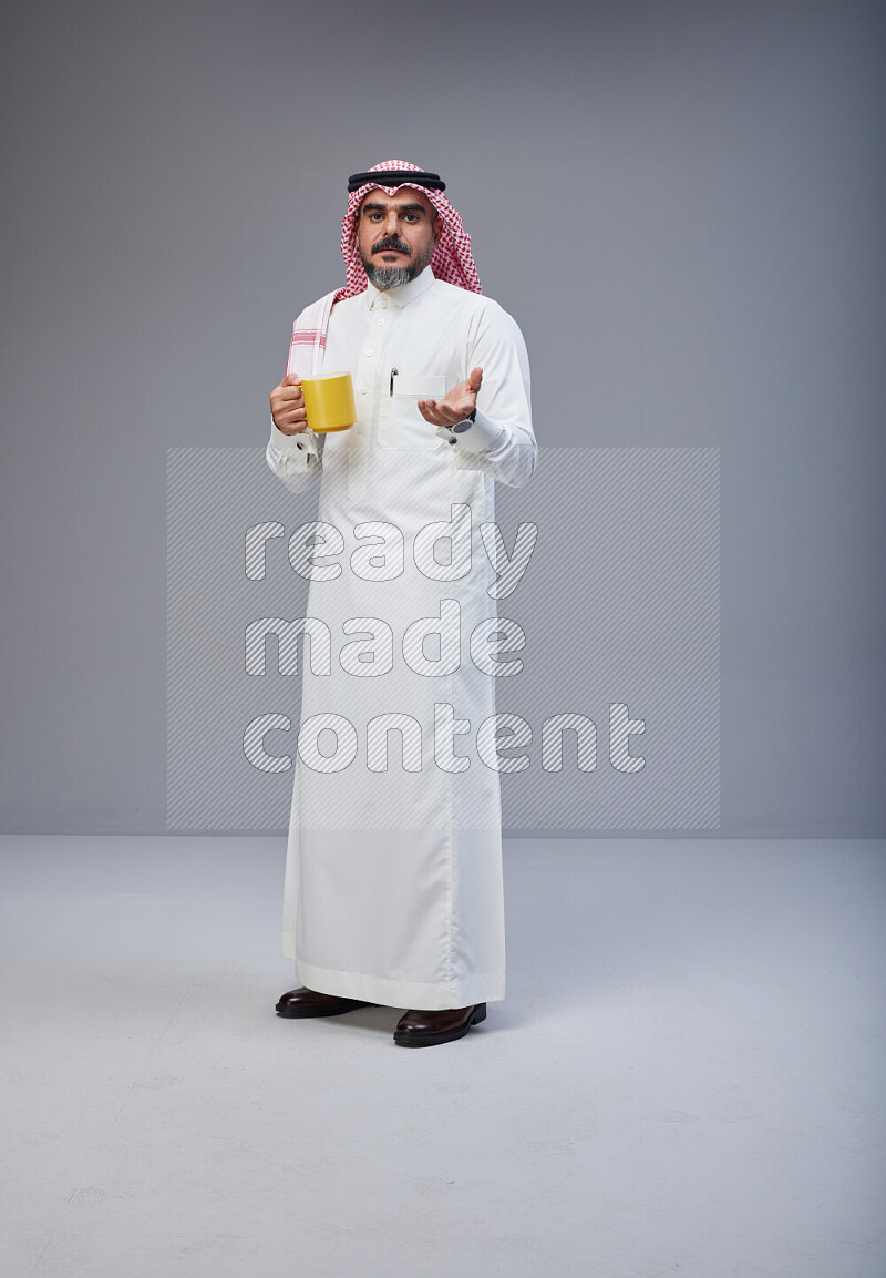 Saudi man Wearing Thob and red Shomag standing holding mug on Gray background