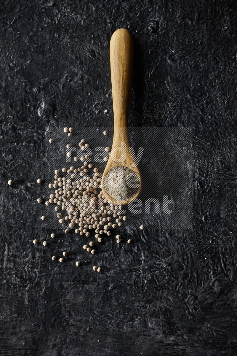 A wooden spoon full of white pepper powder with white pepper beads on textured black flooring