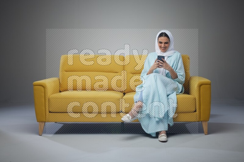 A Saudi woman wearing a light blue Abaya and white head scarf sitting on a yellow sofa and using her phone eye level on a grey background