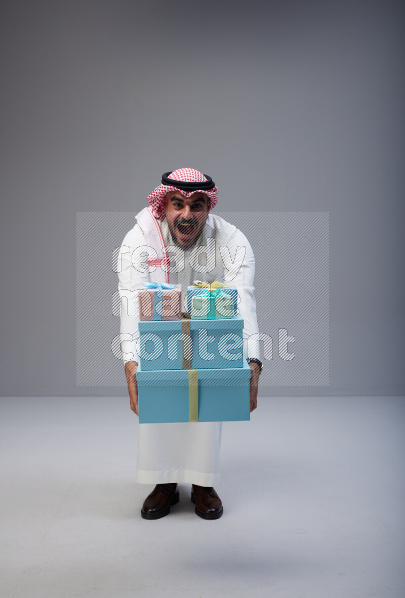 Saudi man Wearing Thob and red Shomag standing holding gift box on Gray background