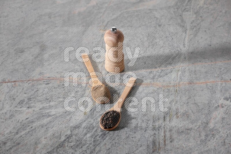 ملعقتان خشبيتان، ملعقة ممتلئة بمسحوق الفلفل الأسود والأخرى ممتلئة بحبات الفلفل الأسود ومطحنة خشبية على خلفية رخام رمادية