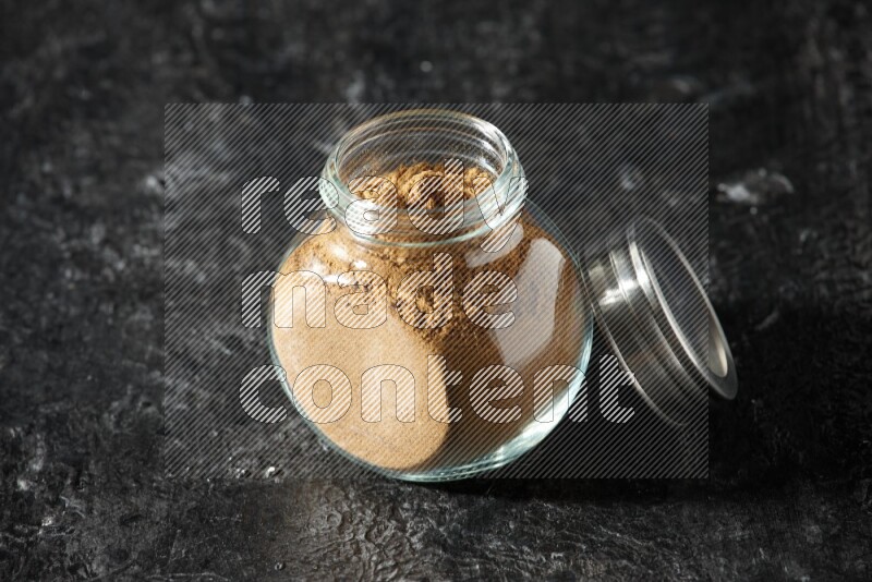 A glass spice jar full of allspice powder on a textured black flooring