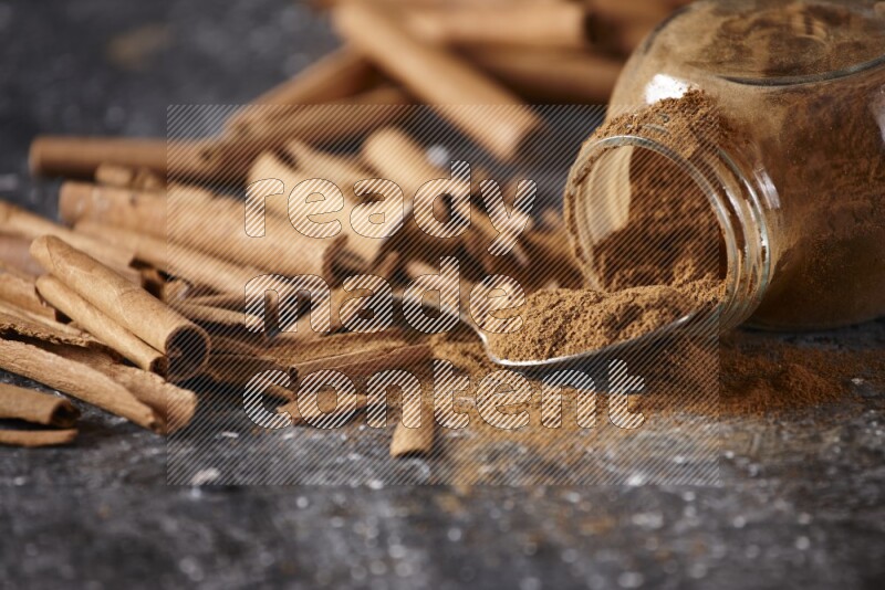Herbal glass jar full cinnamon powder flipped and a metal spoon full of powder surrounded by cinnamon sticks on textured black background in different angles