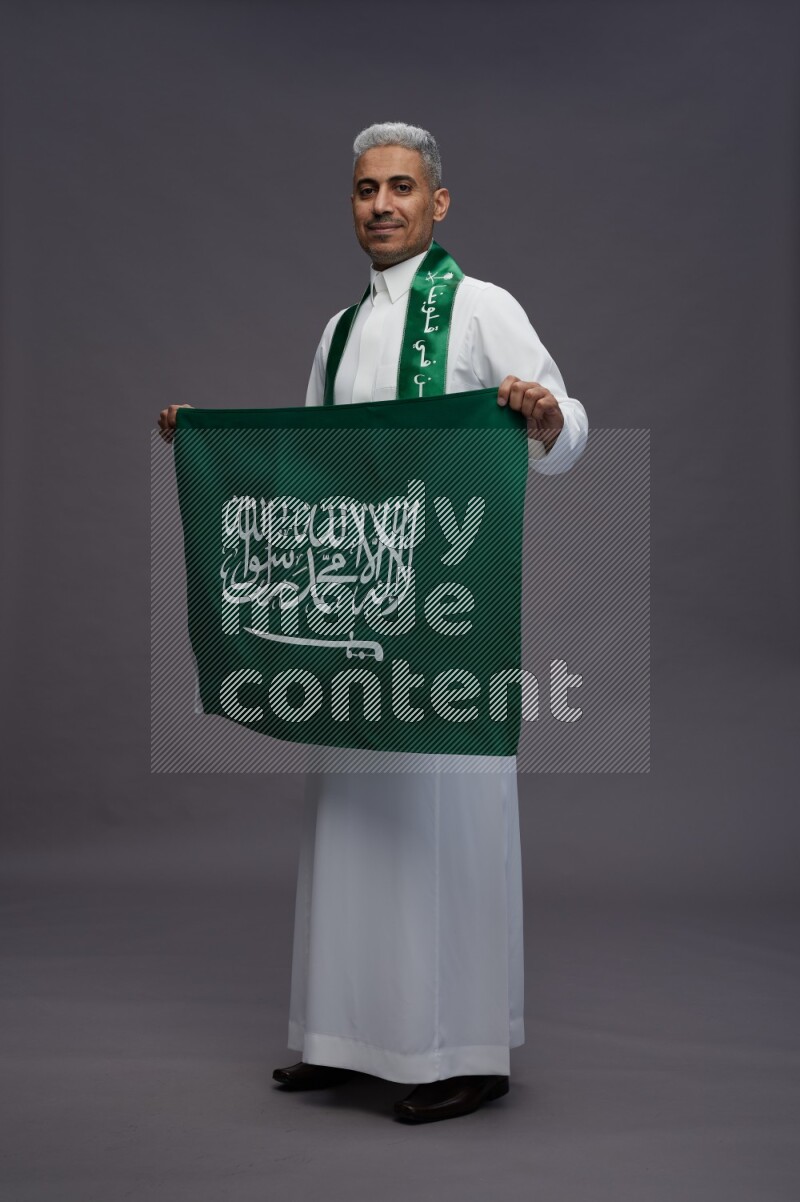 Saudi man wearing thob standing holding Saudi flag on gray background