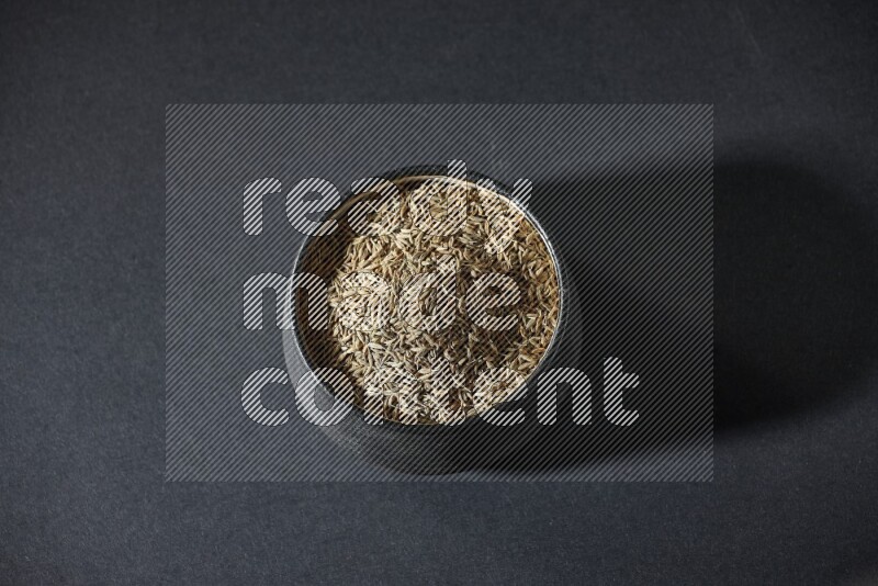 A black pottery bowl full of cumin seeds on a black flooring