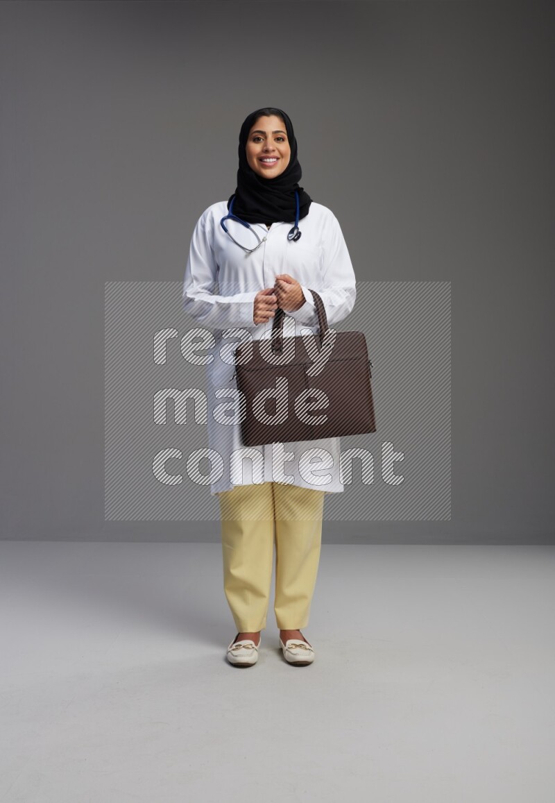 Saudi woman wearing lab coat with stethoscope standing holding bag on Gray background