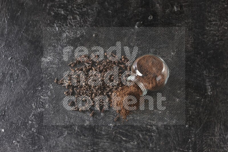A flipped glass spice jar full of cloves powder and powder came out of it with cloves spread on textured black flooring