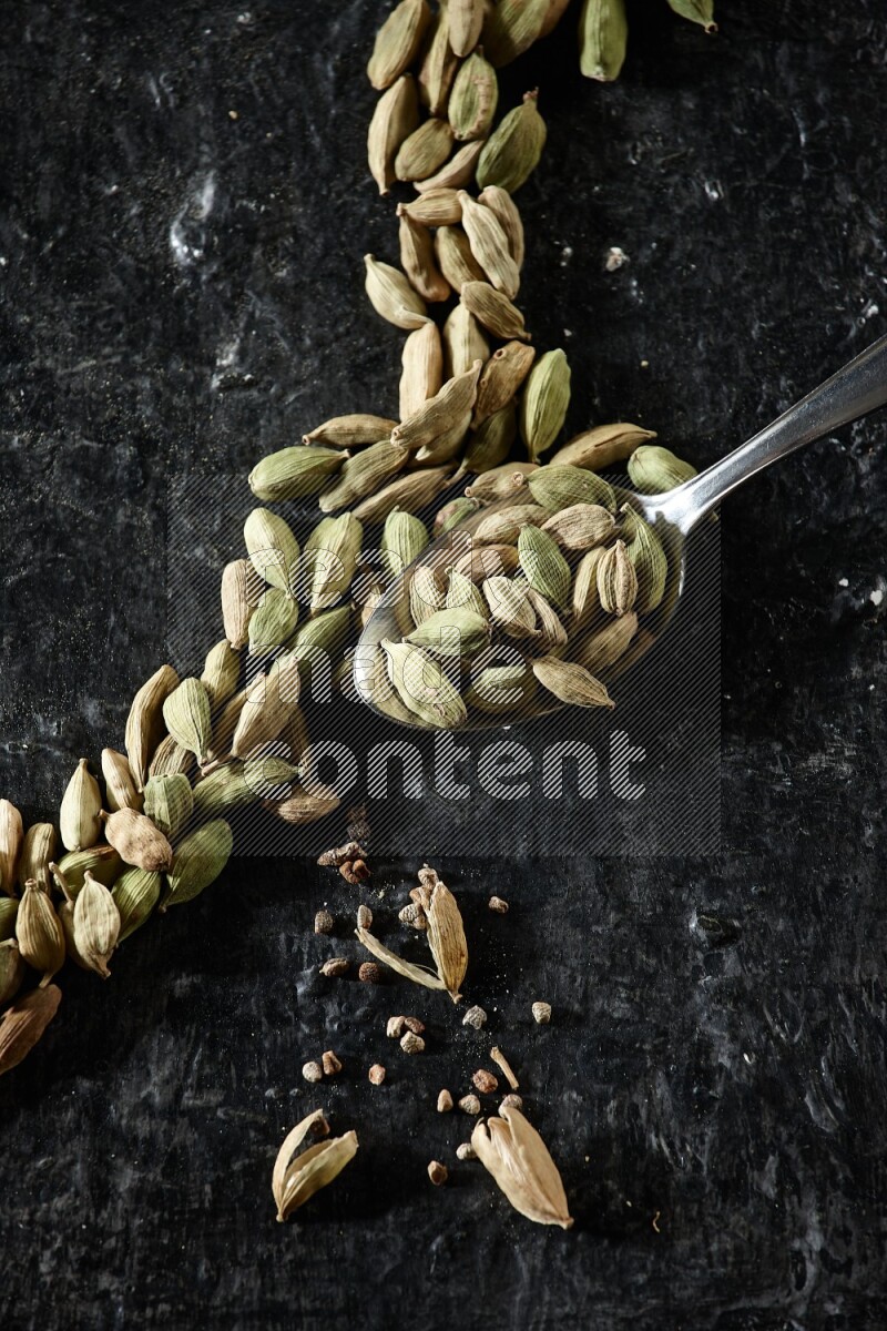 A Metal spoon full of cardamom seeds and some seeds beside it on a textured black flooring