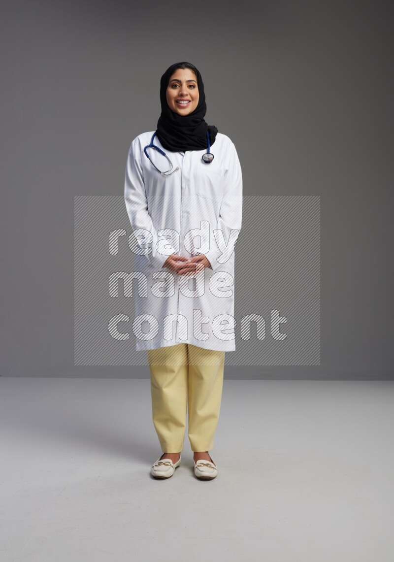 Saudi woman wearing lab coat with stethoscope standing interacting with the camera on Gray background