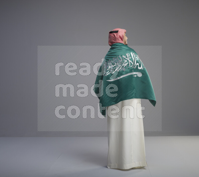 A Saudi man standing wearing thob and red shomag wrapping big saudi flag on gray background
