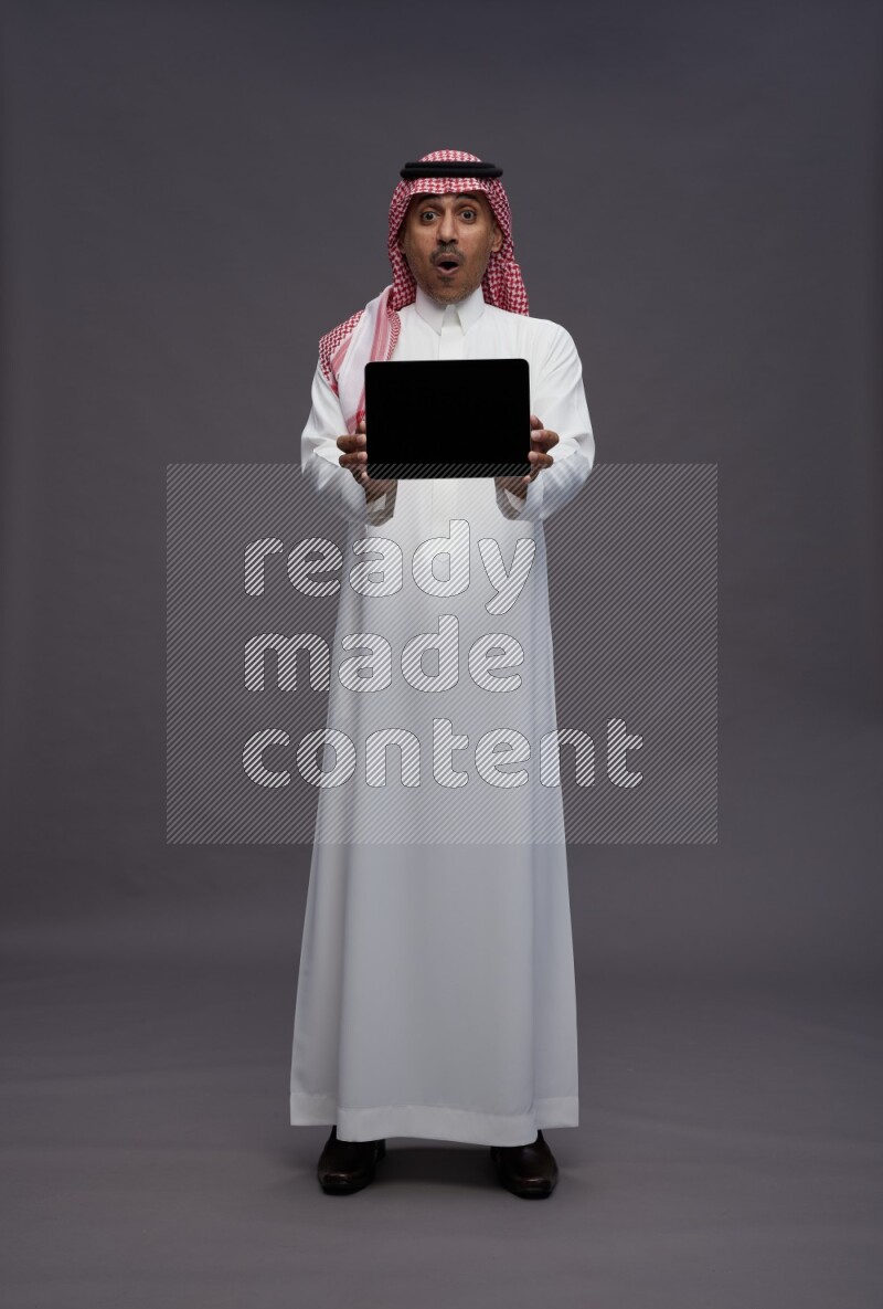 Saudi man wearing thob and shomag standing showing tablet to camera on gray background