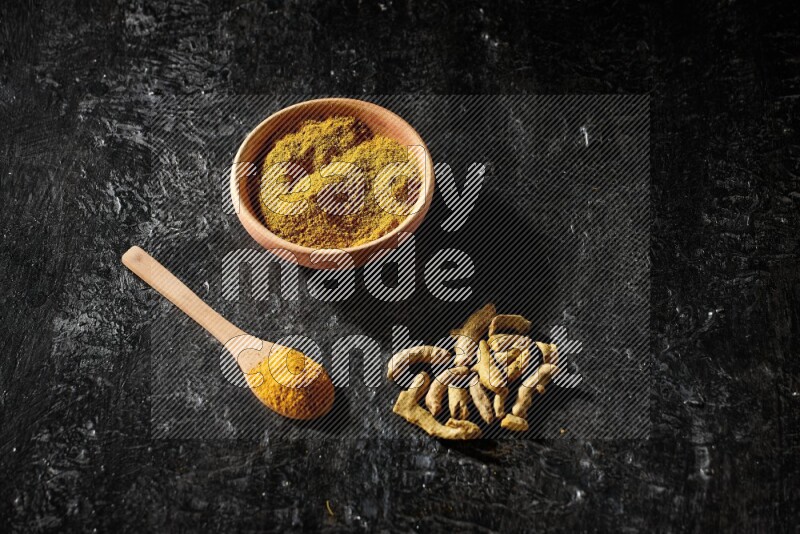 A wooden bowl and a wooden spoon full of turmeric powder with dried turmeric fingers on textured black flooring