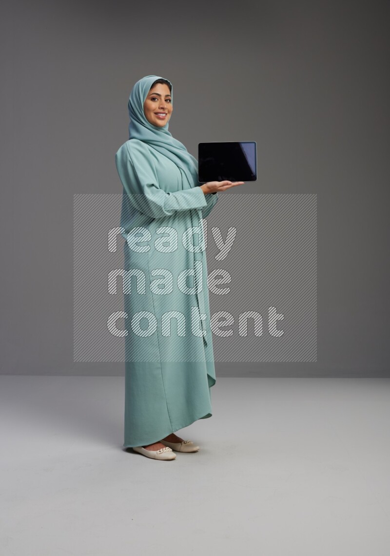 Saudi Woman wearing Abaya standing showing tablet to camera on Gray background