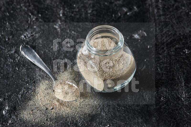 Herbal glass jar and metal spoon full of white pepper powder on textured black flooring