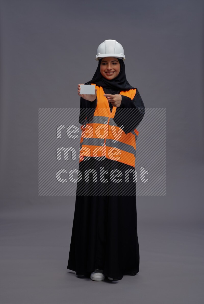 Saudi woman wearing Abaya with engineer vest standing holding ATM card on gray background