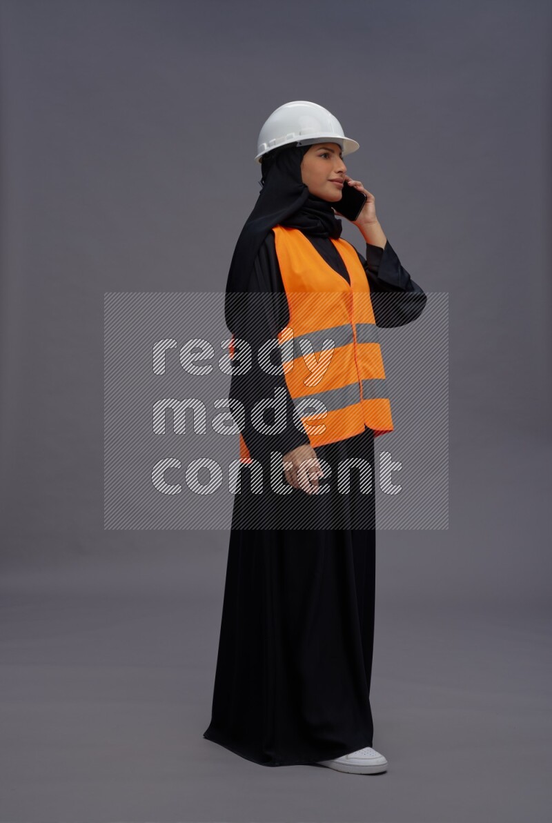 Saudi woman wearing Abaya with engineer vest standing talking on phone on gray background