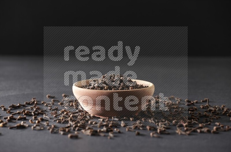 A wooden bowl full of cloves with spreaded whole cloves on a black flooring
