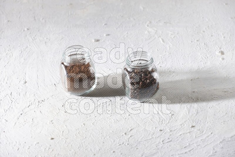 2 glass jars full of cloves powder and cloves grains on a textured white flooring