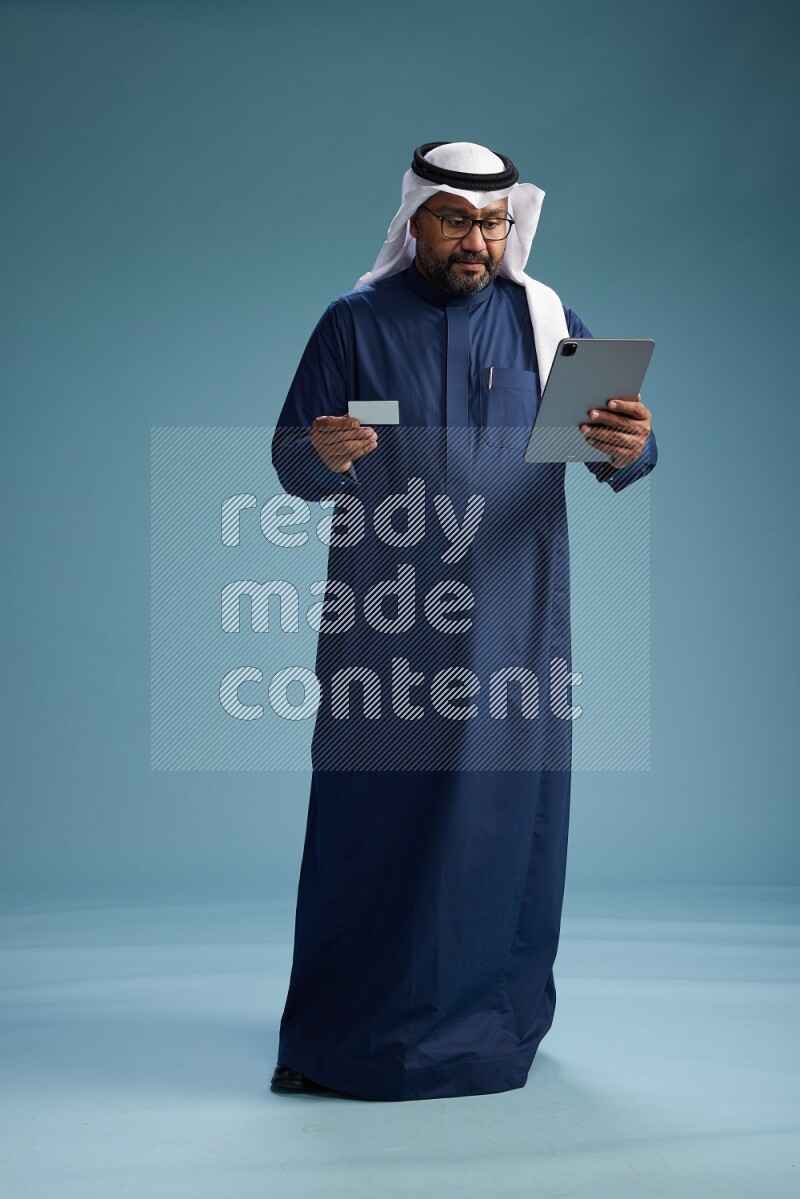 Saudi Man with shimag Standing holding ATM card while working on tablet on blue background
