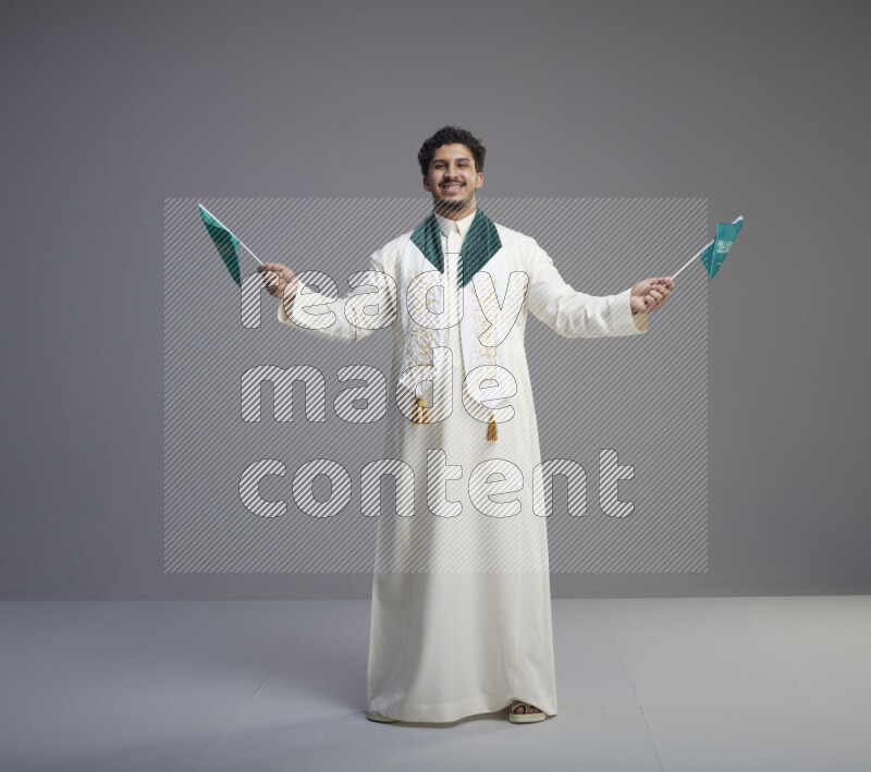 A Saudi man standing wearing thob and saudi flag scarf and holding small saudi flag on gray background