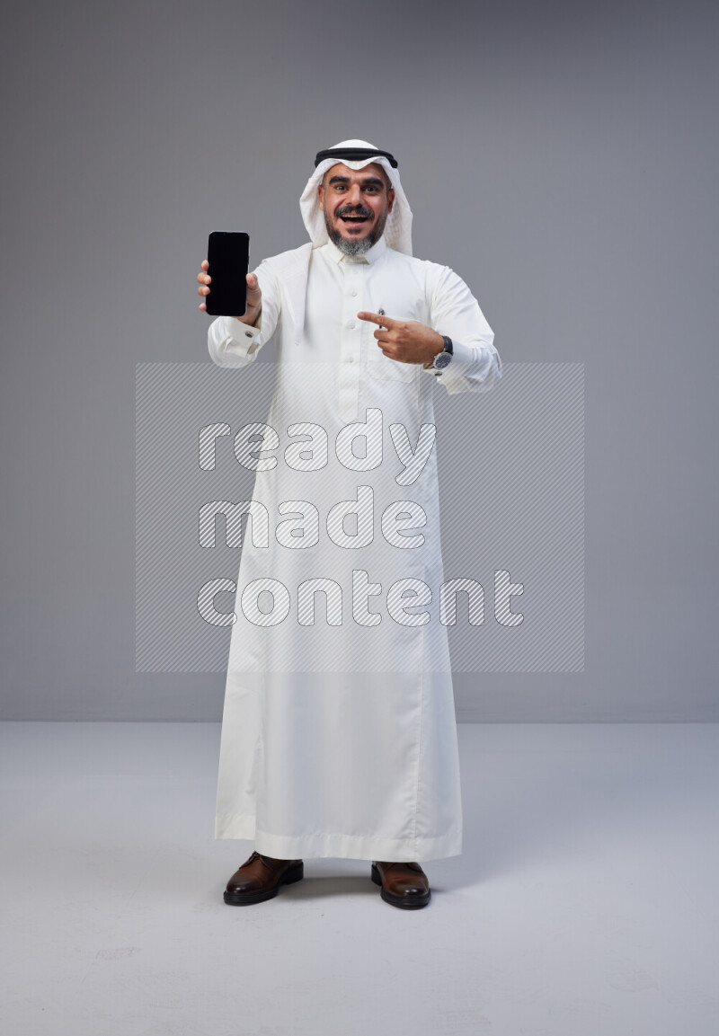 Saudi man Wearing Thob and white Shomag standing showing phone to camera on Gray background
