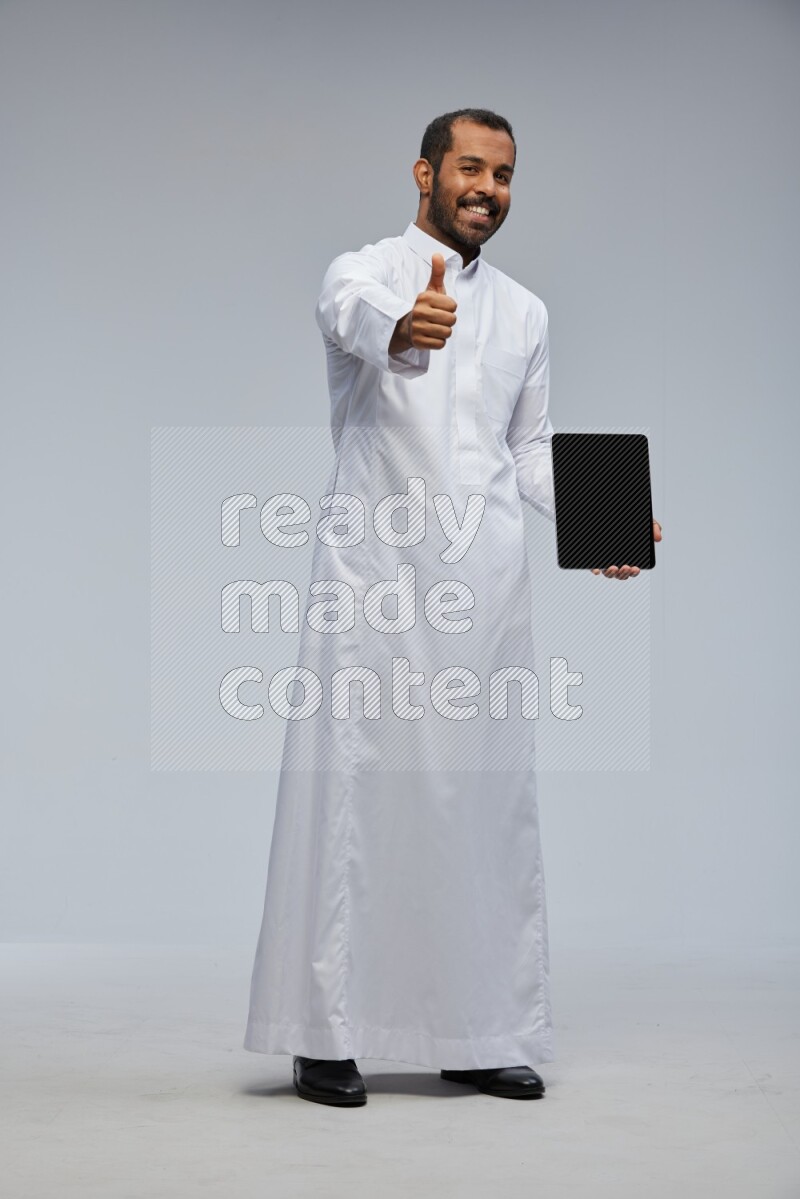 Saudi man wearing Thob standing showing tablet to camera on Gray background