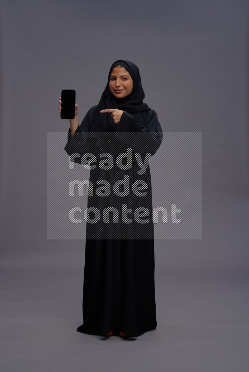 Saudi woman wearing Abaya standing showing phone to camera on gray background