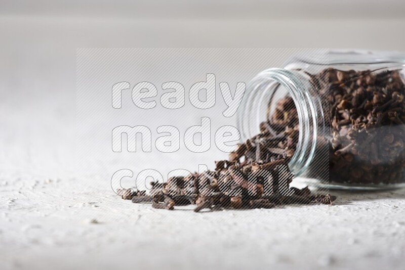 A flipped glass spice jar full of cloves and cloves came out of it on textured white flooring