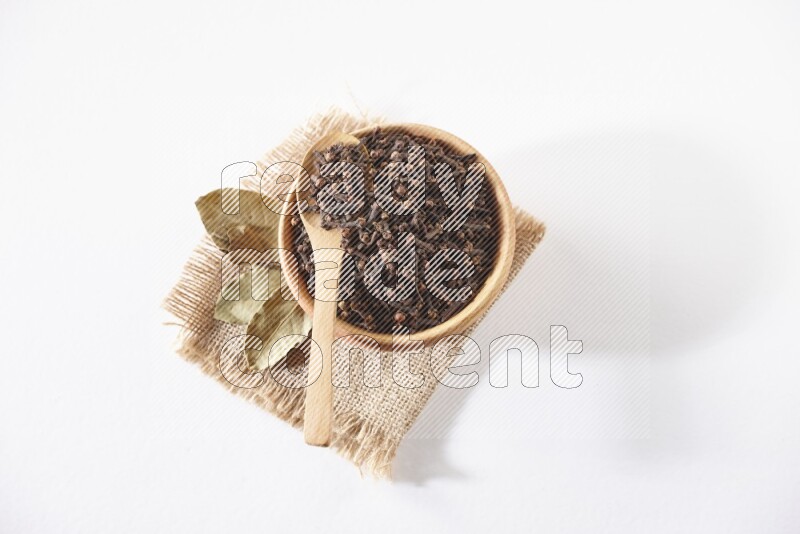 A wooden bowl and a wooden spoon full of cloves on a piece of burlap on a white flooring