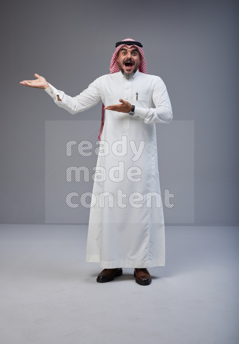 Saudi man Wearing Thob and red white Shomag standing interacting with the camera on Gray background