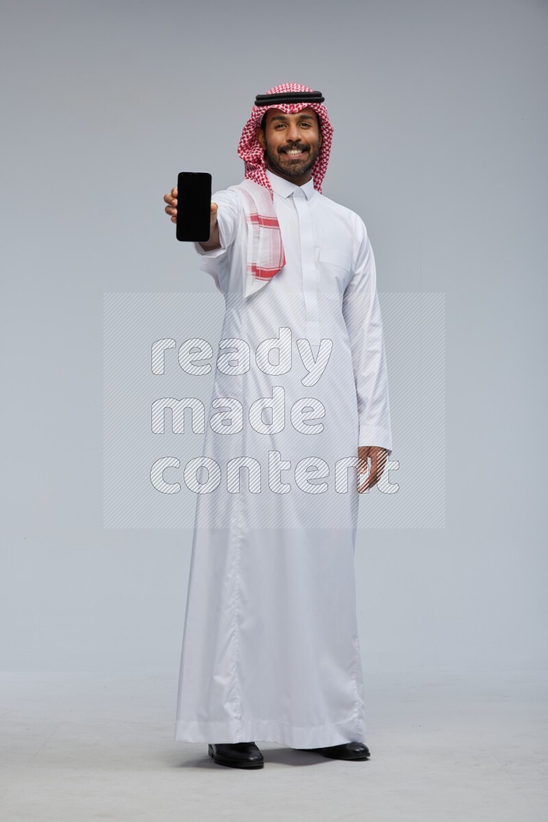 Saudi man Wearing Thob and shomag standing showing phone to camera on Gray background