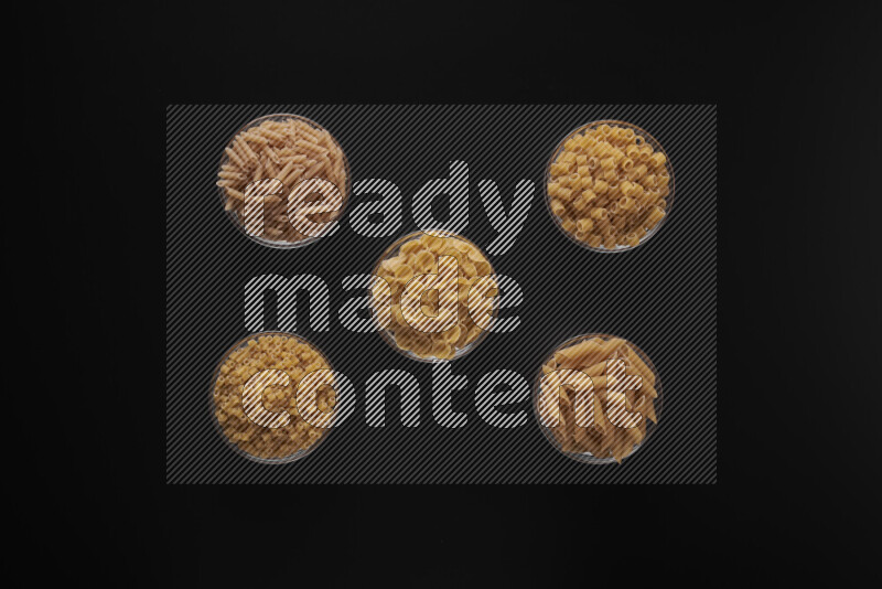 Different pasta types in 5 glass bowls on black background