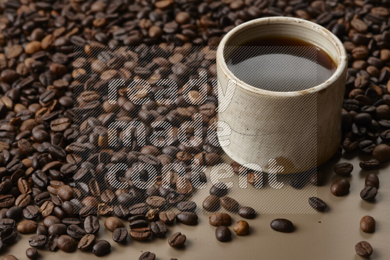 A beige pottery cup of coffee surrounded by roasted coffee beans on beige background
