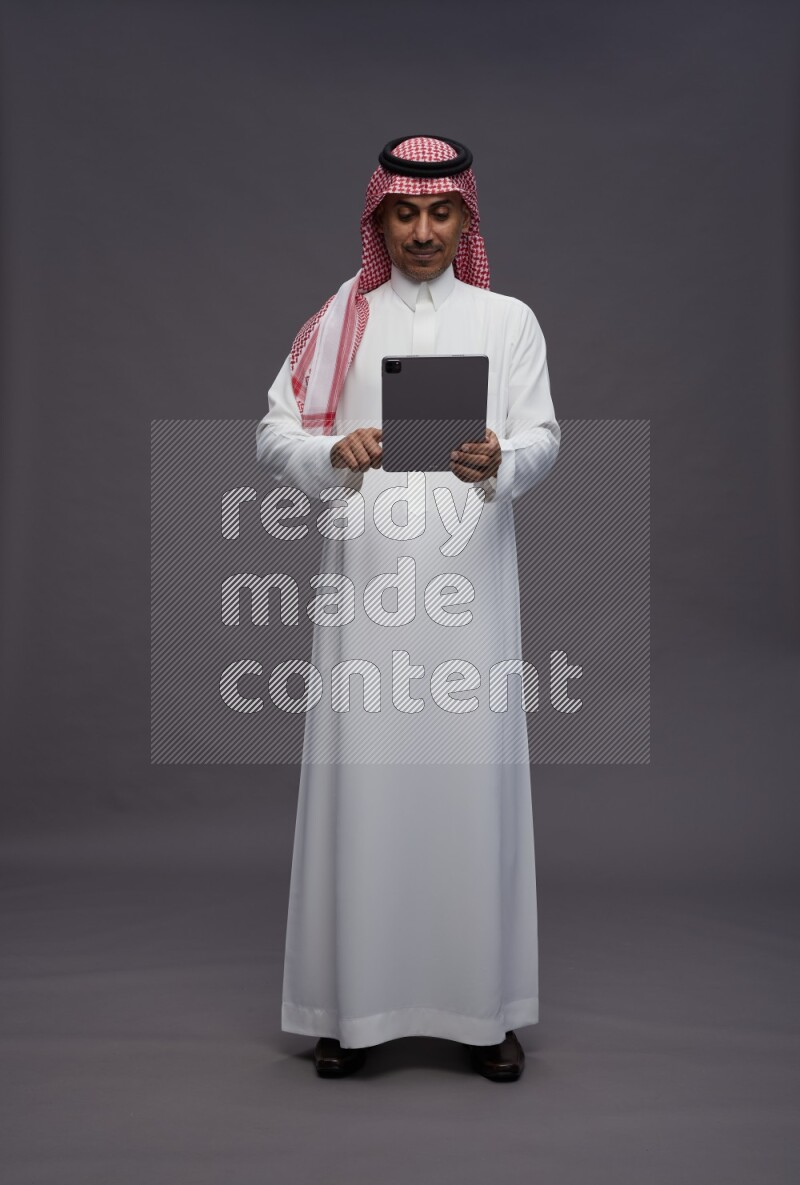 Saudi man wearing thob and shomag standing working on tablet on gray background