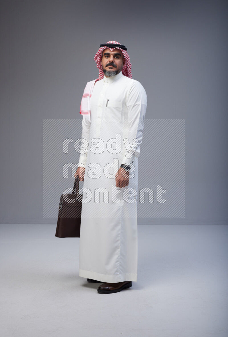 Saudi man Wearing Thob and red Shomag standing holding bag on Gray background