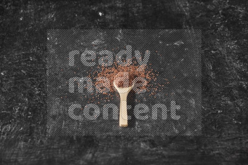 A wooden spoon full of garden cress seeds surrounded by the seeds on a textured black flooring