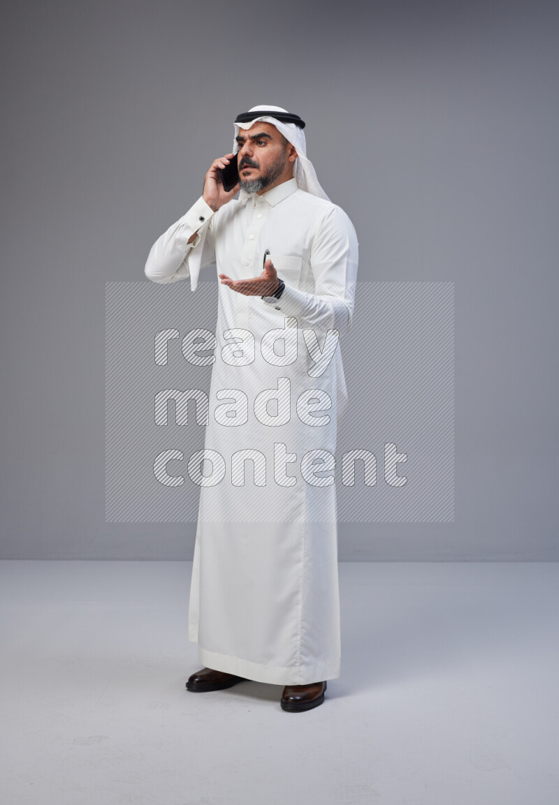 Saudi man Wearing Thob and white Shomag standing talking on phone on Gray background