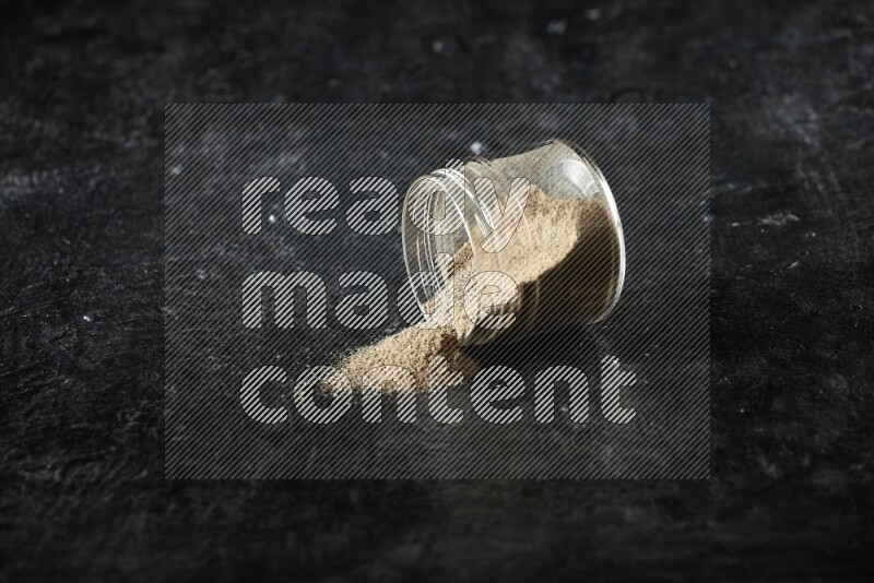 A flipped glass jar full of cardamom powder and powder spilled out of it on textured black flooring