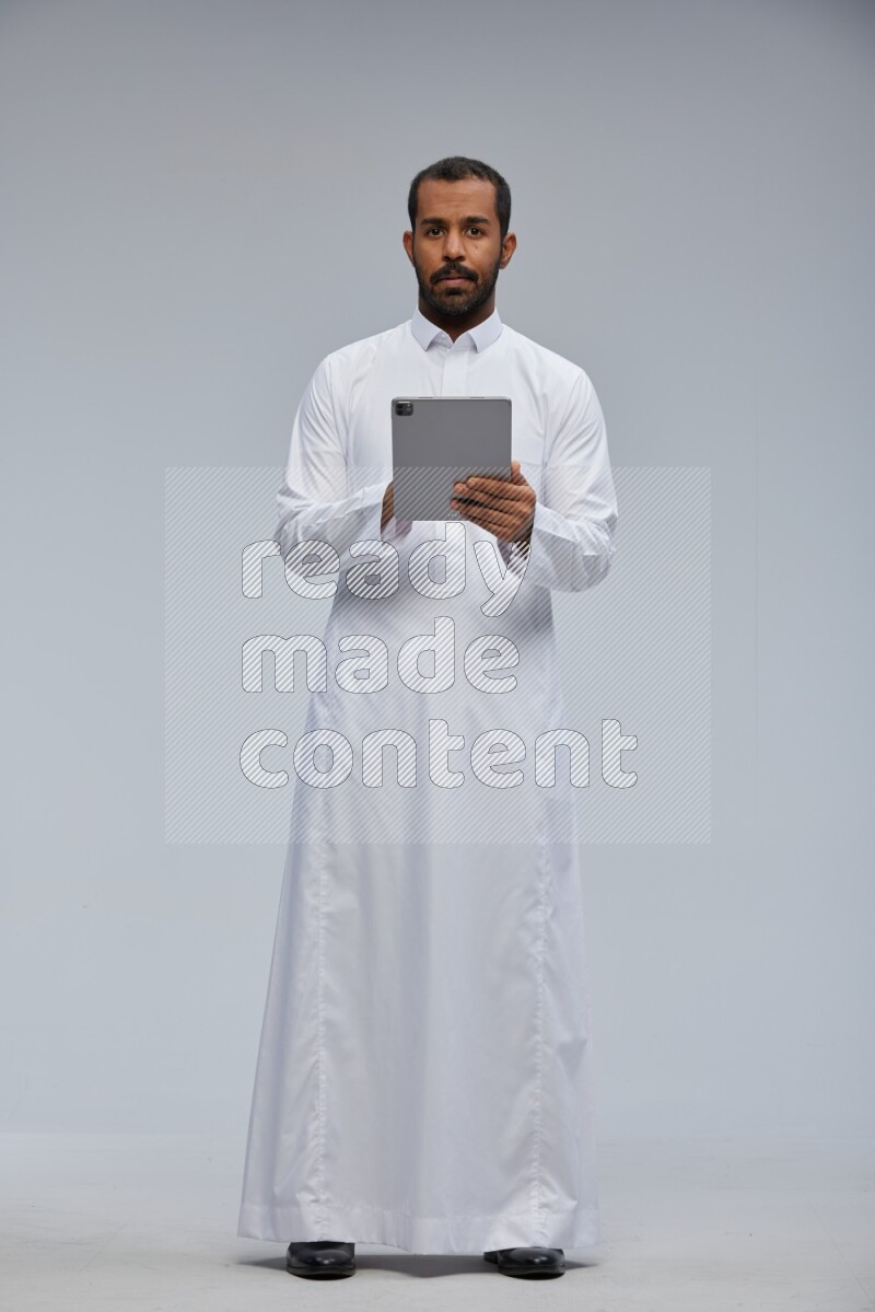 Saudi man wearing Thob standing working on tablet on Gray background